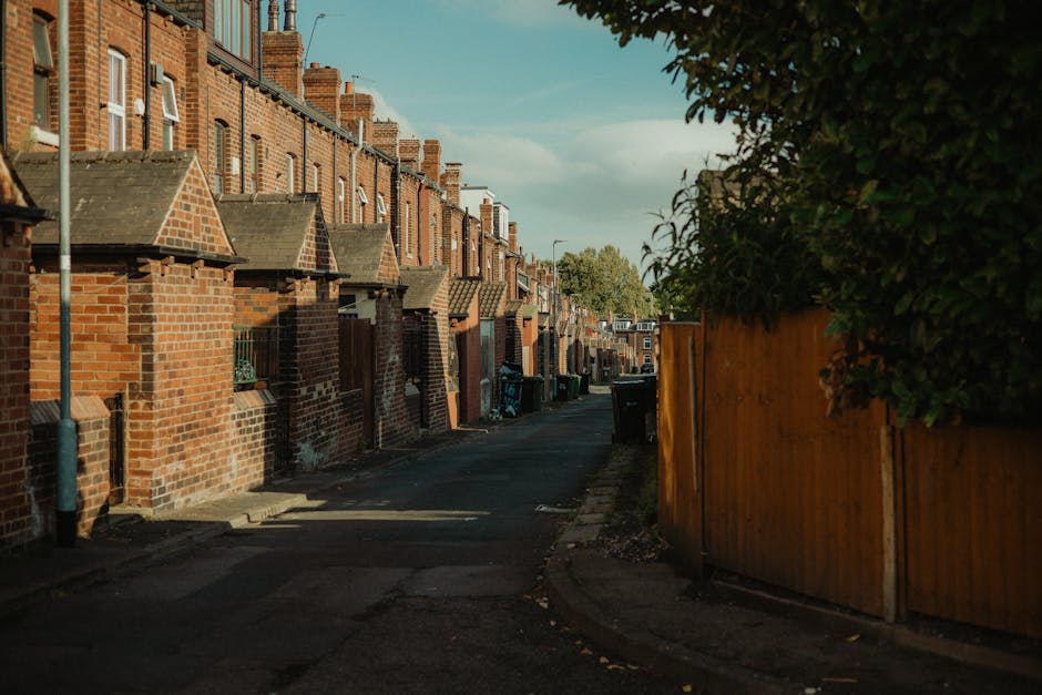 A narrow residential street lined with red-brick terraced houses featuring small front porches and tiled roofs. The lower portion of each house has brick walls, some with small windows protected by metal bars. The street surface is asphalt with visible patches and slight unevenness. On the right side, a tall wooden fence painted in a warm, natural tone partitions the properties from a garden area with dense green foliage extending above the fence. Several black wheelie bins are positioned along the pavement near the houses, likely used for waste collection, and appear to be waiting for collection or disposal. A gray metal pole stands on the left side of the street, possibly supporting street signage or lighting. The scene is illuminated by natural daylight, with soft shadows cast across the street, indicating a partly cloudy sky. The environment and visible objects suggest a typical urban or suburban setting where private waste disposal and rubbish removal services, such as those offered by Waste Collection Harrow, are utilized for managing household waste and ensuring driveway access remains unobstructed for collection vehicles.