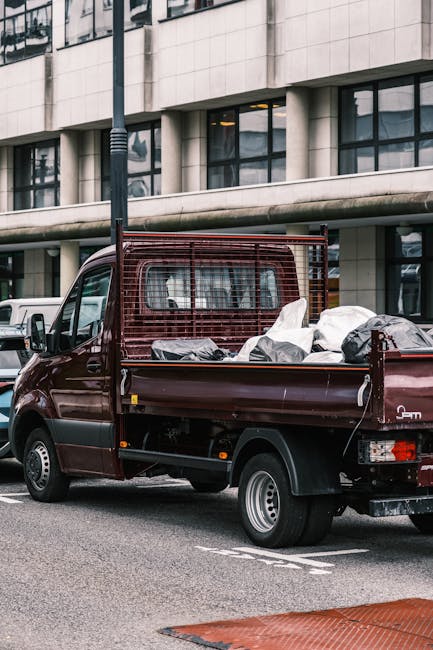 A dark red flatbed mini truck parked on a city street in front of a modern office building with large glass windows and concrete paneling. The truck's cargo bed is filled with various types of rubbish, including white and black plastic bags, some tied and others open, revealing construction debris or miscellaneous waste. The truck has a metal mesh side panel and a tailgate, with one of the bags partially hanging over the edge. Surrounding vehicles are visible nearby, and a lamppost stands close to the truck's front. The environment appears to be an urban area with pavement markings and a designated parking space, suggesting a scene where private or alternative waste collection services, such as those provided by Waste Collection Harrow, are involved in on-site clearance of rubbish not collected through standard municipal services.