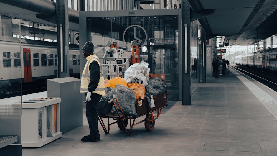 At a modern train station platform, a worker dressed in a black jacket, dark trousers, white gloves, a grey beanie, and a reflective safety vest stands next to a red hand truck loaded with various garbage bags and discarded items. The bags are made of plastic, differing in color from transparent to black and orange, and appear filled with waste or unwanted belongings, some with visible crumples and creases in the material. The background features sleek, glass paneling, metallic structural elements, and a partially visible glass enclosure behind the worker, with mechanical waste processing equipment or sorting machinery behind it. On the left, a large white waste disposal container with a lid is placed on the platform near the worker, while in the distance, additional travelers are visible, along with railway tracks extending into the horizon, under an overhanging metal canopy. The overall setting emphasizes the importance of efficient rubbish and waste management, paralleling private rubbish collection services like Waste Collection Harrow, and highlighting the scene's integration within an infrastructure supporting alternative waste handling.