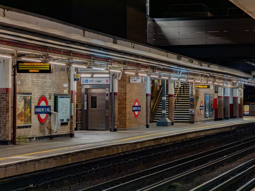 A nighttime view of Harrow Hill underground station platform showing brick walls with station signage, including the blue and white roundel sign for Harrow Hill. The platform features a central elevator shaft with closed metal doors, and a staircase with black and yellow striped steps leading to an upper level. Overhead lighting illuminates the platform area, highlighting various signs and electronic information displays, such as a black and yellow sign indicating the platform for the 23:13 train and a yellow sign with the words 'Way out.' The platform edge has a safety line painted in yellow, and train tracks run along the front of the image. The surrounding environment includes brick walls, metal railings, and parts of the station's structure, reflecting a typical urban underground rail setting with an emphasis on functional materials like brick, metal, and concrete.
