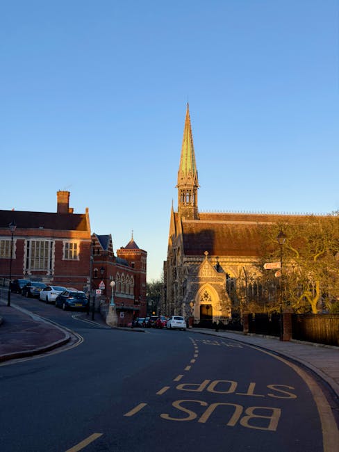 The image depicts a quiet, curved street in a historic area during daylight hours, with several parked cars lining the left side on a paved driveway. Prominently featured is a large medieval-style stone church with a tall, pointed spire and Gothic architectural elements, such as arched windows and detailed stonework, situated on the right side of the street. The church's exterior shows weathered stone in shades of grey and brown, with some moss or lichen growth visible on the surface. To the left of the church, there are red-brick buildings with gabled roofs and chimney stacks, typical of traditional UK architecture. The street surface includes a faded yellow road marking indicating “BUS STOP,” and there is a visible “STOP” sign at the intersection. The background sky is clear with a soft blue tone, suggesting early morning or late afternoon light. The scene is calm, with minimal activity, and gives an impression of an area where private or independent waste collection services, such as those provided by Waste Collection Harrow, could be employed for on-site rubbish removal, especially in the context of managing bulky or construction waste in historic districts.