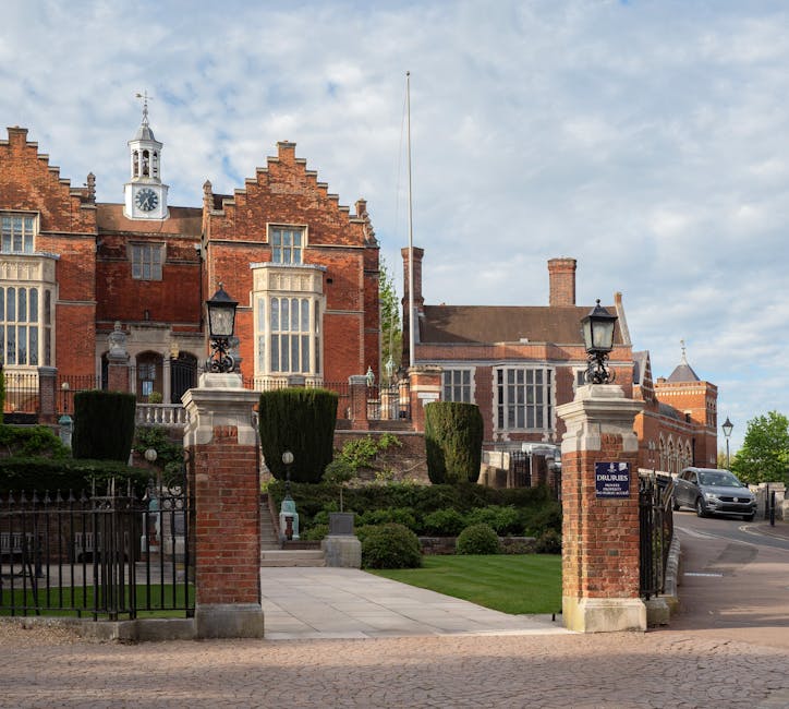 A large historic brick building with a distinctive stepped gabled roof and a central clock tower topped with a weather vane is visible in the background, set behind a gated entrance. The entrance is marked by brick pillars with decorative stone caps and ornate black lamppost-style lamps on top, positioned on either side of the gate. In front of the building, there is a well-maintained garden area featuring neatly trimmed, tall, rectangular box hedges, rounded shrubs, and lush green grass. A paved pathway leads from the gate into the garden, and a street with parked cars runs to the right of the scene. The sky overhead is partly cloudy, providing natural light that softly illuminates the scene. The overall setting suggests a prestigious or historic property, which may require regular rubbish removal or private waste management, supported by local waste collection services such as Waste Collection Harrow, to maintain its tidy appearance.
