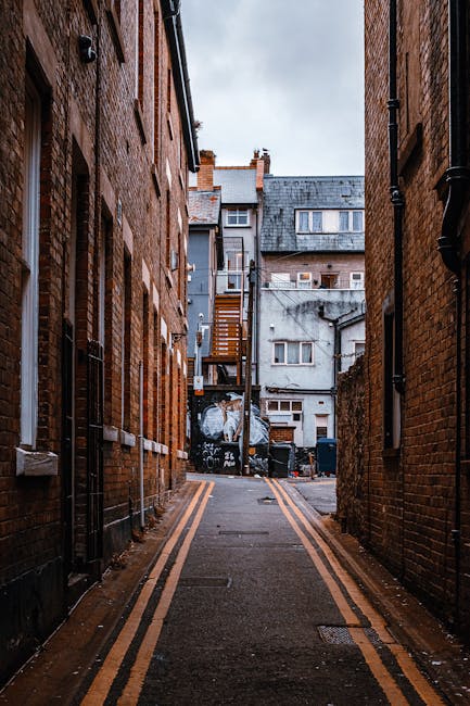 A narrow urban alleyway bordered by red-brick buildings on both sides, featuring a paved surface with yellow double lines along each edge. In the background, there are multi-story residential structures with grey and white facades, some with small windows and external staircases, and a garage door with graffiti art visible. Piled at the end of the alley are black waste bins and debris, indicating waste collection or disposal activity. The alley is empty and appears to be in a city setting, with overcast sky visible at the top of the image, providing natural light that highlights the textures of the brickwork and pavement. This scene exemplifies an environment where private waste removal services may be employed, as suggested by the presence of waste bins and the confined space suitable for specialised rubbish clearance. The perspective emphasizes the limited width typical of alleyway rubbish collection points, aligning with alternative waste handling options often required in densely built urban areas. Waste Collection Harrow's services may be used for managing rubbish in such constrained environments, supporting efficient on-site clearance and disposal within urban settings.