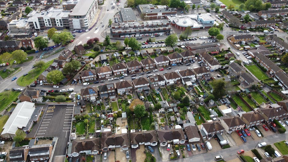 An aerial view of a suburban residential area with rows of terraced houses featuring slate tiled roofs and small front gardens, some with patio furniture and lawn areas. Several vehicles, including cars and vans, are parked along the narrow streets and driveways. In the foreground, a large parking lot adjacent to a community center or commercial building is visible, with clearly marked spaces and a few parked cars. Behind the houses, there is a mixture of apartment blocks and commercial buildings, some with flat roofs and modern facades, set amidst trees and greenery. The environment appears calm and orderly, with well-maintained properties and a mix of green lawns, paved pavements, and landscaped gardens, consistent with a typical UK suburb. Waste Collection Harrow's rubbish services are relevant to this scene, where private waste disposal and household rubbish collection are often managed through local or independent collection providers, subtly reflected by the organized layout suited for waste removal activities.
