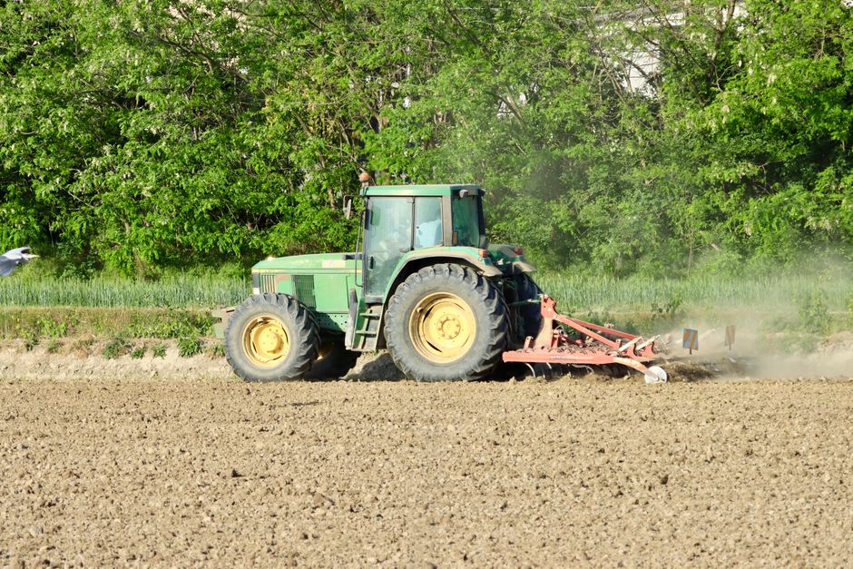 A green tractor with yellow wheels is operating on a large, flat, freshly tilled field with a textured soil surface, creating a cloud of dust and loose dirt as it moves across the land. The tractor features a enclosed cab with clear glass windows, and its front-mounted implement is actively working the soil, possibly for preparation or planting. Behind the tractor, there is a dense row of leafy green trees with vibrant foliage, indicating a rural or agricultural environment widely associated with outdoor waste management and land clearing activities. The scene is illuminated by natural daylight, with subtle shadows cast by the tractor and surrounding trees, emphasizing the clarity and detail of the soil and equipment. The overall setting reflects typical agricultural practices, aligning with the context of independent or private site clearance often requiring rubbish removal services that specialize in handling soil, vegetation, and farm waste, as provided by Waste Collection Harrow.