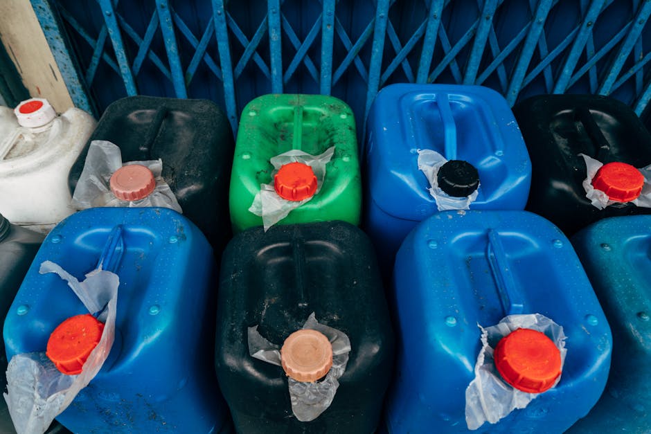 A collection of plastic jerry cans arranged outdoors, sitting on a paved surface in front of a blue metal fence. The jerry cans are in various colors including black, blue, green, and white, with some showing signs of dirt and weathering. Each container features a large, circular screw-on cap in contrasting colors such as red, black, and beige, with some caps wrapped partially in frayed, transparent plastic bags. The containers are positioned close together, with some leaning slightly, and are likely used for storing or transporting liquids. The background consists of the blue fencing, indicating an industrial or storage area suitable for waste or chemical container collection. The overall scene is well-lit, with natural daylight highlighting the textures and colors of the containers, aligning with services provided by Waste Collection Harrow for waste or hazardous material disposal, emphasizing the importance of proper handling and private waste management options.